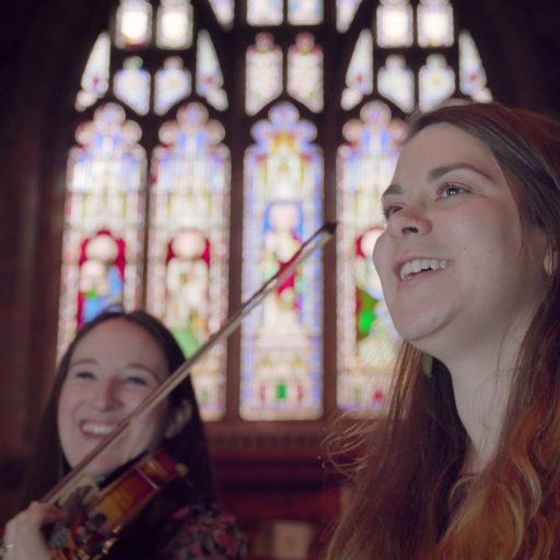 A violinist and singer in a church, with a large stained glass window in the background.