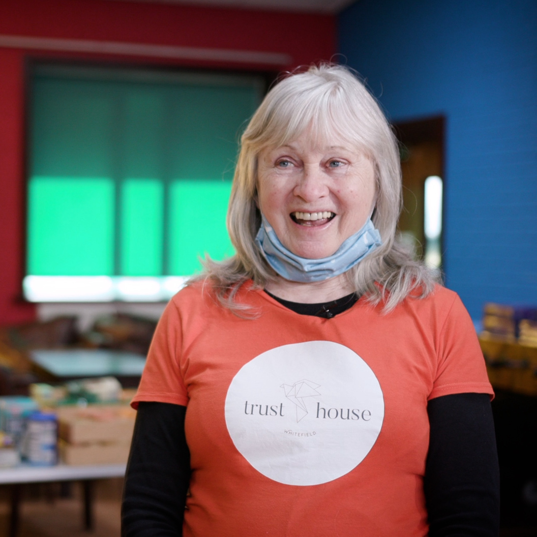 A woman smiling in a community centre, wearing a t-shirt with the Trust House logo