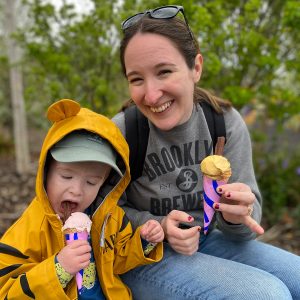 A boy and a woman eating ice cream from cones. The boy is wearing a yellow rain jacket with animal ears, and the woman is wearing a grey jumper.