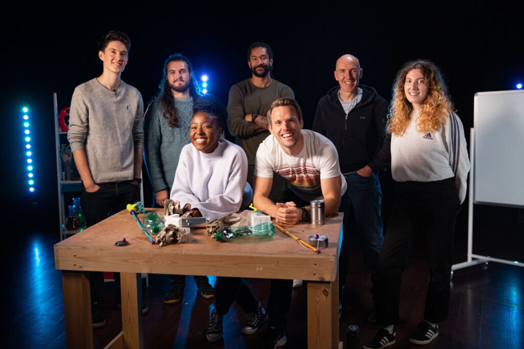 Cast and crew of BBC Teach's 'Show Me the Science'. Two presenters smiling with a TV production crew behind them, in a dark studio set with bright blue lights.