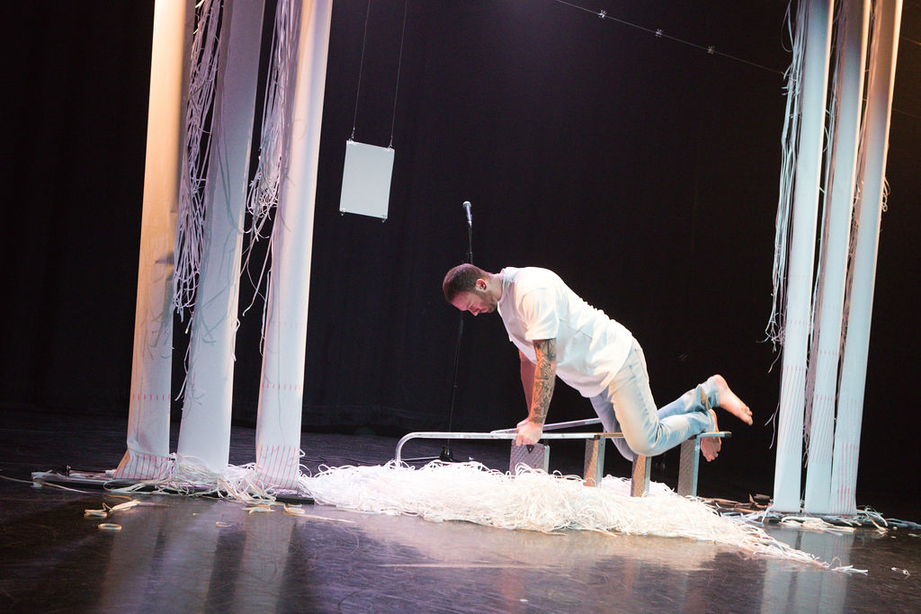 Photo of a theatre performance. A man holds himself up in a plank position on a horizontal ladder. He is surrounded by shredded paper, and more reams of paper are hanging down from above him, some of them partly shredded.
