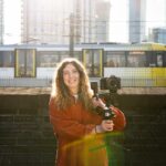 Woman in a red shirt holding a camera on a gimbal stabilising device, with a Manchester Metrolink tram in the background, and tall glass buildings behind that.