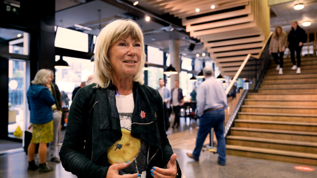 Screengrab of an interview clip with a woman in the foyer of an arts venue, with other conference delegates milling around behind