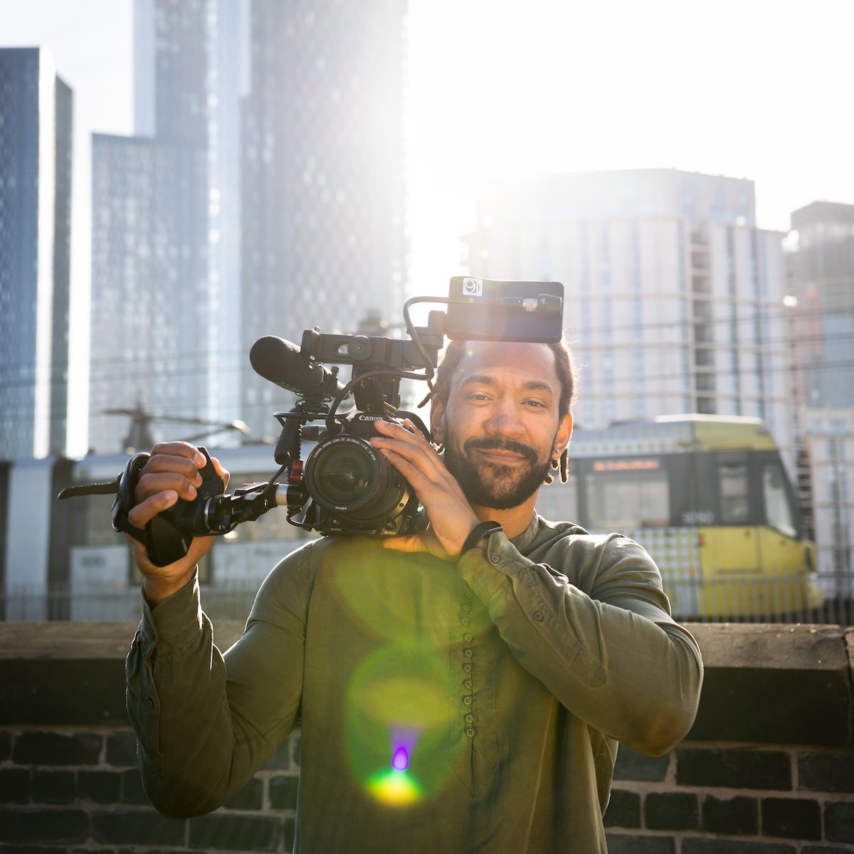 Jamie Swaby holding a video camera to his shoulder, with a tram and Deansgate Square towers in the background