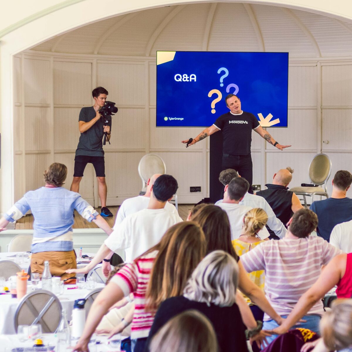Corporate event with a man on stage leading a room full of people in a stretch. A slide on the screen behind him says Q&A. Next to the screen is Ben Horrigan with a video camera on a gimbal, filming the event.