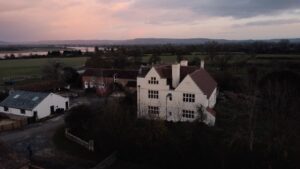 Drone shot of a large farmhouse building surrounded by farmland, with a sunset in the background